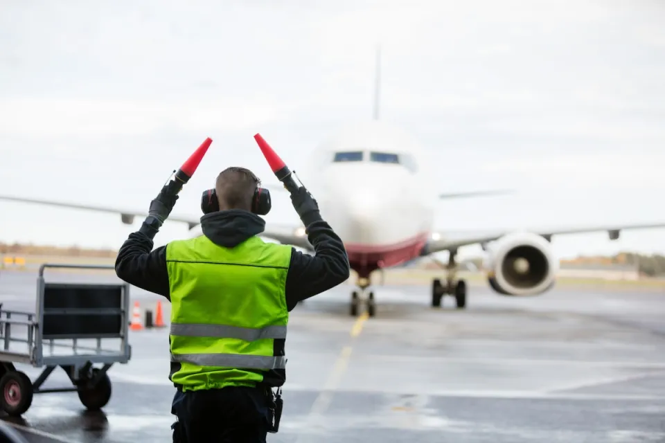 Ground crew person signaling to airplane with red batons on runway on a rainy day.